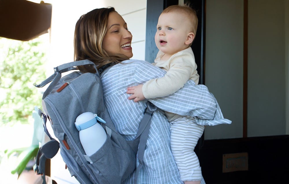 A woman holding a baby has a portable bottle warmer in the side pocket of her backpack