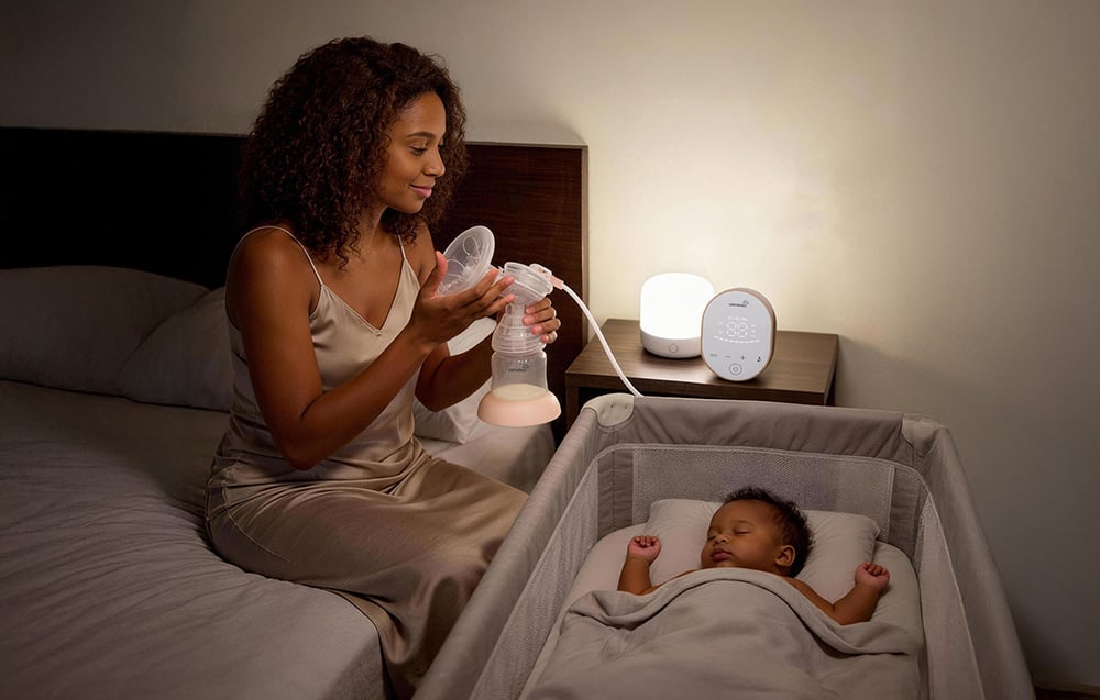 A woman is sitting by the bed using a breast pump while her baby sleeps in the crib next to her