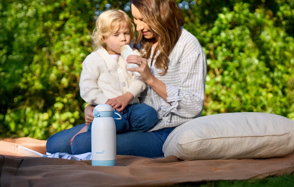 A woman feeds her baby on a blanket in the garden, with a portable bottle warmer standing next to her