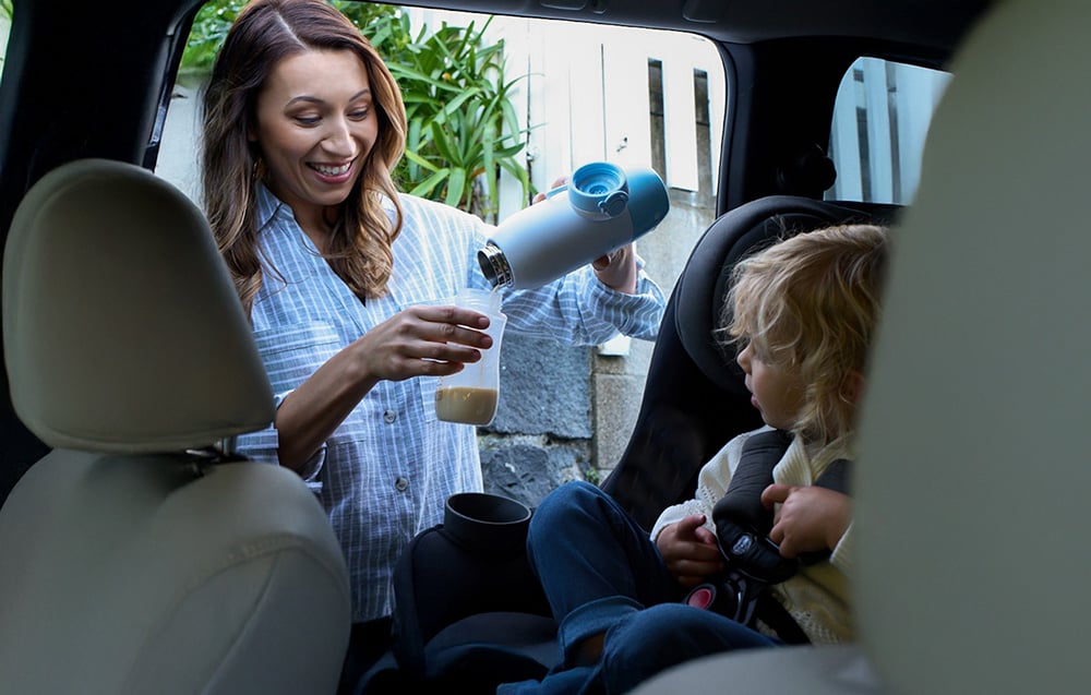 A woman in a car pours milk from a portable bottle warmer into a bottle, while her baby sits in a car seat