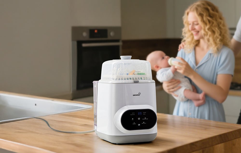 Multifunctional bottle washer operating on a kitchen countertop, with a woman feeding a baby in the background