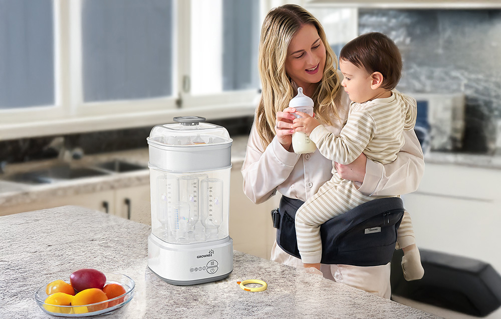 Woman holding a child at the kitchen counter, sterilizer with bottles standing next to her