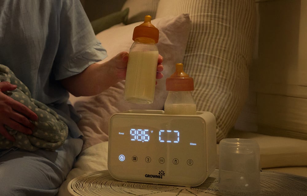 A woman sits with her baby by the bed and holds a bottle next to the warmer with an illuminated display