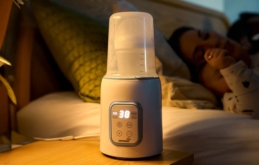 Bottle warmer on a bedside table, next to a sleeping baby with mother in the background