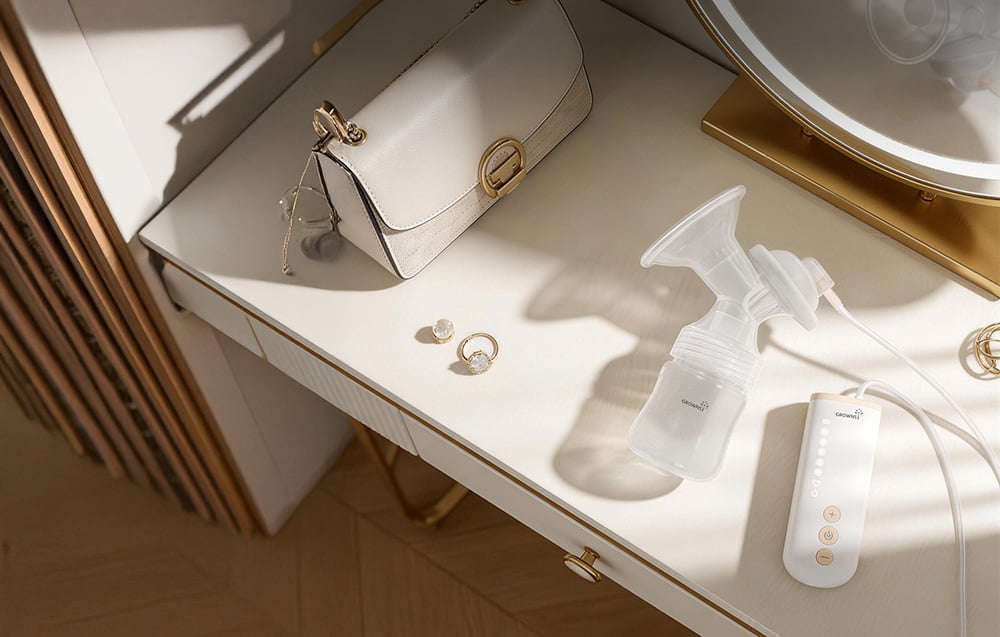 An electric breast pump with a bottle and control panel lies on a light-colored dressing table next to a handbag and jewelry