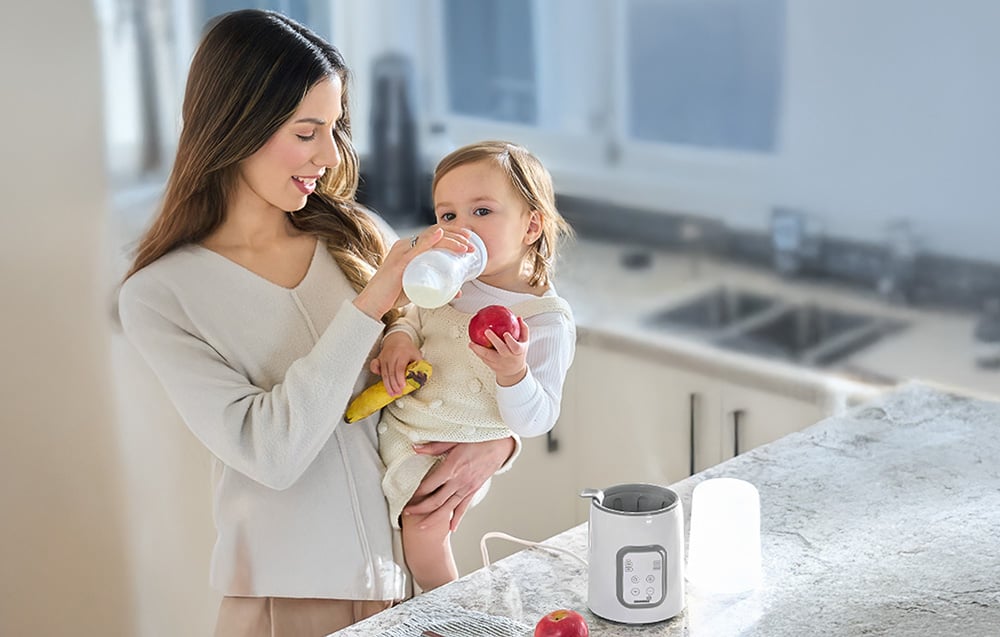 A woman feeds her baby with a bottle in the kitchen, with a bottle warmer on the counter next to her