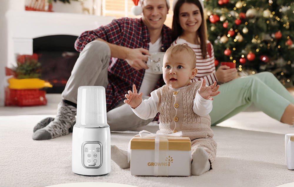 A child sits on a rug next to a bottle warmer and a gift, with parents in the background by the Christmas tree